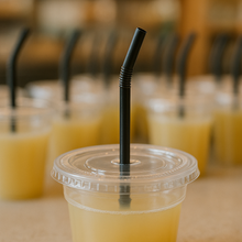 Load image into Gallery viewer, 7.75&quot; long black straw with a flexible next in which the flexible section is 0.5&quot; long, sticking out of the center hole of a clear juice cup lid, on a counter at a juice shop. many other similar juice cups in the background
